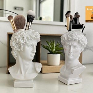 以下が含まれることがあります： 白い胸像型のホルダーが2つあり、それぞれクラシックな彫刻に似ており、メイクブラシが詰められています。 ホルダーは白い表面にあり、背景には小さな植物と木製の箱があります。 装飾品です。
