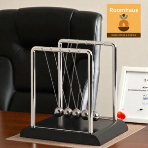 Può includere: Un giocattolo da scrivania a culla di Newton con cinque sfere d'acciaio lucido sospese a un telaio cromato. La base è nera e il giocattolo poggia su una superficie marrone. Il logo Roomhaus è visibile sullo sfondo.