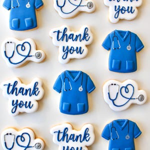 May include: A selection of decorated sugar biscuits shaped like blue medical scrubs, stethoscopes, and the words "thank you" in blue icing. The biscuits are arranged on a white surface.