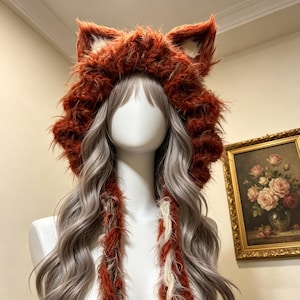 Peut inclure: Une capuche en fausse fourrure orange rouille, dotée d'oreilles d'animal dressées avec des accents intérieurs couleur crème. La capuche possède de longs liens tressés pendants en fourrure orange rouille et crème, portée sur un mannequin aux longs cheveux ondulés.
