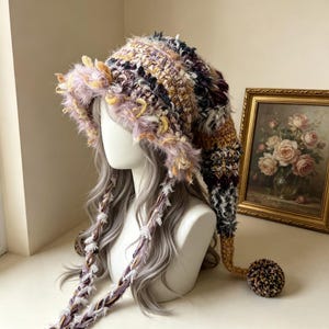 May include: A long, pointed crochet hat featuring a mix of textured yarns in shades of lavender, mustard yellow, dark brown, and cream. The hat has a wide, fuzzy brim, two long braided side ties with frayed edges, and a large pom-pom at the tip.