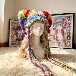 May include: A multi-colored crochet jester hat featuring a vibrant rainbow pattern with two pointed peaks, each topped with a small pom-pom. The hat includes long, dangling braided fringe strands in various colors that extend down from the sides.