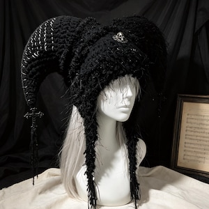 Puede incluir: Un gorro de bufón de ganchillo negro con dos cuernos grandes y curvados decorados con imperdibles plateados. El gorro presenta un pequeño emblema de calavera plateada en el centro y largos flecos negros con colgantes de cruz.
