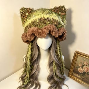 May include: A fuzzy, olive green and lime green knit hat featuring cat-like ears adorned with small brown bows and buttons. The hat has a wide, ruffled brown faux fur brim and long, dangling green yarn fringe accents that hang down the sides.