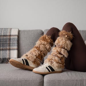 Peut inclure: Une paire de baskets blanches avec des bandes latérales noires, portées avec des jambières en fausse fourrure marron. Les jambières présentent une texture hirsute en couches et sont ornées de petits boutons en bois marron clair.