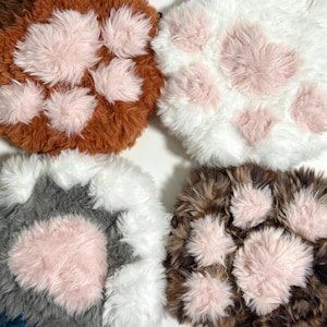 May include: Four plush paw-shaped mats featuring long faux fur in brown, white, grey, and brown-black variegated colours. Each mat has soft, light pink faux fur pads arranged in a paw print pattern, providing a textured and tactile surface.
