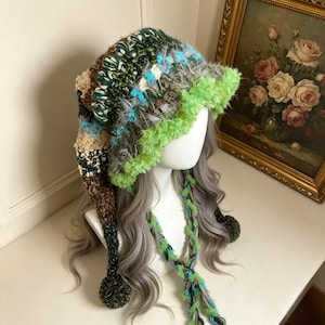 May include: A long, slouchy crochet hat featuring a mix of textures and colours including dark green, brown, tan, and bright neon green. The hat has a fuzzy neon green brim, dangling braided ties with pom-poms, and a long, pointed tail.
