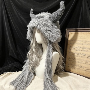 May include: A grey faux fur hat featuring two prominent, upright horns and long, dangling side panels. The hat is crafted from a shaggy, textured material that covers the head and extends down the sides, creating a distinct, horned silhouette.