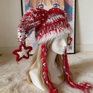May include: A red and white crocheted jester-style hat with two long, pointed ear-like flaps. The hat features a fuzzy, textured yarn finish, small white star-shaped accents, and a large, solid white star dangling from one of the pointed ends.