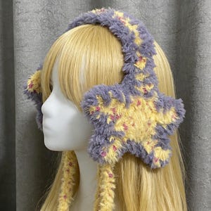 Op de afbeelding: Een pluizige, stervormige oorwarmer in tinten lavendel, geel en roze. De oorwarmers worden gedragen op een mannequinhoofd met blond haar. De oorbedekking is van zacht, fuzzy materiaal.