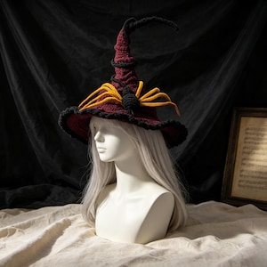 May include: A dark maroon crocheted witch hat featuring a black brim edge and a large, textured black and yellow spider decoration attached to the front. The hat has a tall, pointed crown and is displayed on a white mannequin head with long hair.