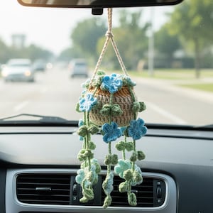 May include: A crocheted hanging planter with blue and green floral details, suspended from a car's rearview mirror. The planter is made with beige, brown, and green yarn, creating a textured, decorative item. The design includes hanging vines with flowers.