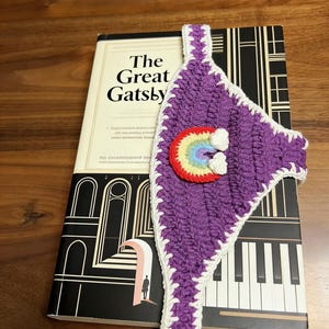 Könnte beinhalten: Ein lila Häkel-Lesezeichen mit Regenbogen- und Wolken-Design, platziert auf einer Ausgabe von "The Great Gatsby". Das Lesezeichen hat einen weißen Rand und besteht aus strukturiertem Garn. Das Buchcover zeigt eine Schwarz-Weiß-Illustration.