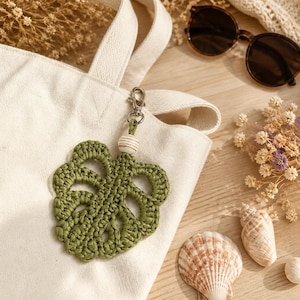 May include: A green, crocheted monstera leaf keychain with a wooden bead accent and silver-tone clasp. The keychain hangs from a white canvas tote bag. Sunglasses, seashells, and dried flowers are also visible.