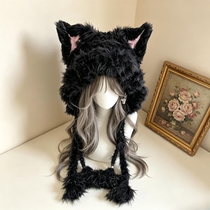 May include: A black faux fur hat featuring prominent cat ears with light pink inner accents. The hat has a thick, shaggy texture and includes two long, dangling earflaps that hang down from the sides, each finished with a fluffy pom-pom end.