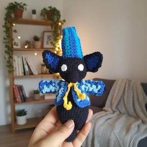 May include: A handmade crochet doll in the shape of a bat, featuring a black body, large blue ears, and a blue and turquoise jacket. The doll has a tall blue hat and a yellow scarf. White circles are used for the eyes.