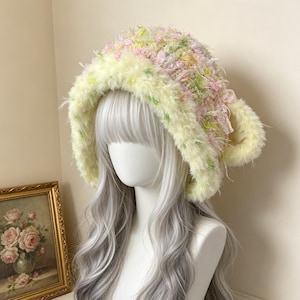 May include: A fuzzy bucket hat featuring a pastel color palette of pink, yellow, and green yarn. The hat has a thick, textured brim in a pale yellow shade and is decorated with a small pink ribbon bow on the side, adding detail to the knit.
