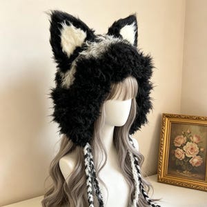 May include: A fluffy black and white faux fur hat featuring prominent animal-like ears on top. The hat has a thick, textured pile and includes two long, braided tassels hanging down from the sides, crafted from a mix of black and white fibers.