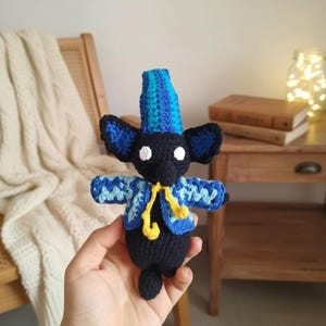 May include: A handmade black crochet doll with large blue ears and a tall blue and turquoise hat. The doll wears a blue and white jacket with a yellow scarf. The doll is being held in a person's hand.
