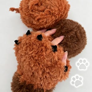 Op de afbeelding: Vingerloze handschoenen gemaakt van pluizig, gestructureerd roestoranje garen met donkerbruine accenten aan de basis en kleine, zwarte, driehoekige stoffen stekels langs de bovenrand. De handschoenen hebben een zachte, pluche uitstraling met twee witte pootafdrukken.