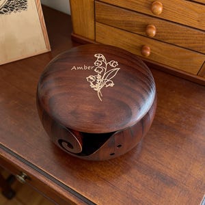 May include: A dark brown wooden yarn bowl with a lid, engraved with the name "Amber" and a floral design. The bowl has a curved slot for yarn. It sits on a wooden table. A wooden drawer unit is in the background.