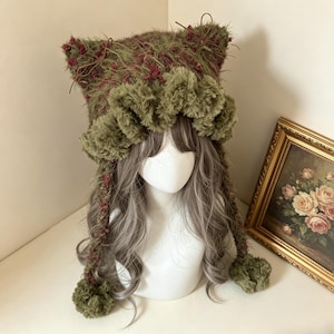 May include: A fuzzy olive green and maroon knit hat featuring cat-like ears, a ruffled brim, and two long braided side ties ending in pom-poms. The hat is displayed on a mannequin head with long wavy grey hair, showcasing a textured yarn construction.