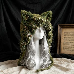 May include: A fuzzy olive green hood featuring prominent cat-like ears and an intricate embellishment of small, dried white flowers woven throughout the textured material. The hood is displayed on a mannequin head with long, wavy silver hair.