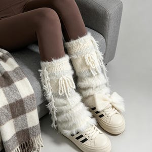 Puede incluir: Un par de calentadores de piernas blancos y esponjosos con textura de punto, lazos decorativos laterales y flecos colgantes, usados sobre zapatillas de deporte color crema con rayas negras laterales. Los calentadores tienen un aspecto suave y peludo con lazos de hilo blanco a juego.