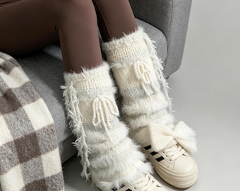 May include: A pair of white fuzzy leg warmers featuring a knit texture, decorative side bows, and dangling fringe accents, worn over cream-coloured trainers with black side stripes. The leg warmers have a soft, shaggy appearance and are tied with matching white yarn bows.
