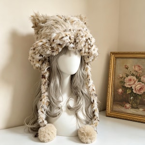 May include: A fluffy, faux fur hat featuring a beige and brown leopard-like spotted pattern. The hat has two pointed ear-like shapes on top and two long, dangling side flaps that end in large, round, solid beige pom-poms made of soft material.