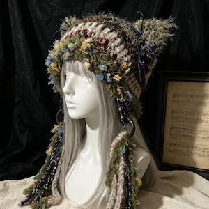 May include: A crochet cat-ear hat featuring a textured mix of olive green, brown, cream, and blue yarn. The hat includes dark blue ribbon bows on the ears and long, dangling yarn tassels attached to large brown rings on each side of the head.