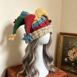 May include: A multi-colored crochet jester hat featuring three long, pointed sections in green, red, and yellow. The hat has a textured, speckled knit base with a ribbed brim and small, star-shaped crochet ornaments dangling from each point.