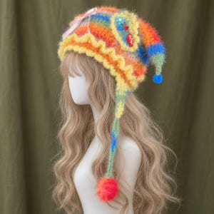 May include: A fuzzy, multi-coloured knit hat featuring a rainbow striped pattern, a yellow ruffled brim, and a light pink ribbon bow. The hat includes a long, dangling side panel with a bright red pom-pom and a small blue pom-pom at the tip.