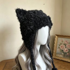 May include: A black fuzzy beanie with pointed cat-ear shapes at the top. The hat is crafted from a textured, shaggy material and features two long, braided ties hanging down from the sides, each finished with a small pom-pom at the end.