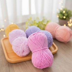 May include: A collection of colorful yarn balls in shades of pink, purple, and yellow, arranged on a wooden tray. The yarn is soft and fluffy, perfect for knitting or crocheting projects. The background includes a window and decorative lights.