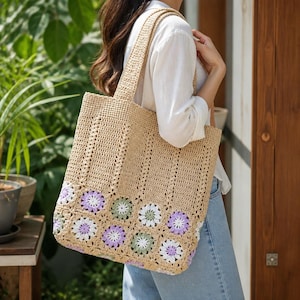 May include: A beige crochet tote bag featuring a vertical ribbed pattern on the upper section and a row of floral motifs in shades of lavender, sage green, and white along the bottom. The bag has two matching crochet shoulder straps.