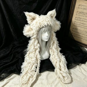 May include: A cream-colored, long-eared animal hat crafted from a combination of chunky cable-knit yarn and shaggy faux fur. The hat features two prominent, pointed ears at the top and two long, dangling side panels that extend downward.