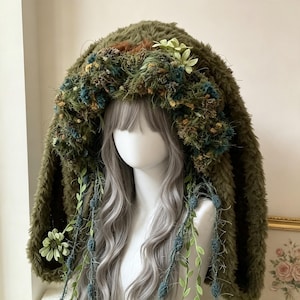 May include: A fuzzy olive green hooded cowl featuring a thick trim of textured moss, dried blue and yellow flowers, and hanging vine-like strands with small green leaves and blue pom-poms, displayed on a mannequin with long wavy gray hair.