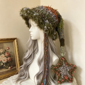 May include: A textured, multi-colored crochet hat featuring a wide, shaggy brim in shades of olive green, brown, and orange. The hat includes long, dangling yarn fringe and a large, five-pointed star ornament attached to a long, trailing tail.