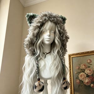 May include: A hooded hat made of long, shaggy faux fur in shades of gray, brown, and green, featuring two pointed animal-like ears on top. Two braided cords hang down from the sides, each ending in a fluffy, leopard-print pom-pom with dark beads.