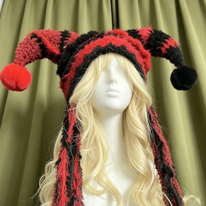 May include: A jester-style hat featuring a fuzzy, textured knit pattern in alternating red and black stripes. The hat has two long, pointed side extensions that terminate in one red pom-pom and one black pom-pom, with long dangling tassels.
