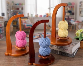 May include: Three wooden yarn holders with different colored yarn. The holders have a curved arch design and a wooden base. One holds pink yarn, another blue, and the third yellow. The yarn is wound into neat balls, perfect for knitting or crocheting projects.