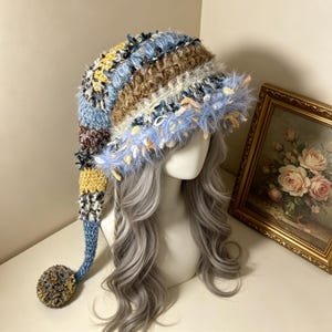 May include: A long, slouchy knit hat featuring a multi-colored striped pattern in shades of blue, yellow, brown, and white. The hat has a textured, fuzzy yarn construction with a wide, frayed brim and a long tail ending in a large, round pom-pom.