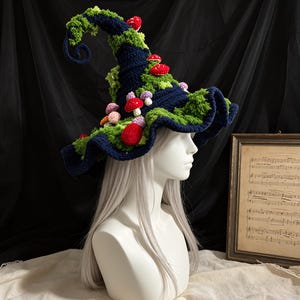 Puede incluir: Un sombrero de ganchillo azul oscuro con una punta alta y curva y un ala ancha y ondulada. El sombrero está decorado con hilo verde brillante texturizado similar al musgo y varios pequeños hongos de ganchillo en rojo, rosa y morado.
