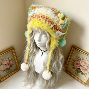 May include: A fuzzy, multi-colored knit hat featuring horizontal stripes of orange, white, and yellow. The hat has long braided earflaps ending in white pom-poms, decorated with small felt shapes including a bone, wings, and a blue butterfly.