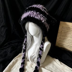 Può includere: Un berretto in lana soffice con strisce orizzontali nere e color lavanda. Il cappello presenta due protuberanze a forma di orecchie sulla parte superiore e due lunghe nappe intrecciate che pendono dai lati, ciascuna terminante con un pompon nero.