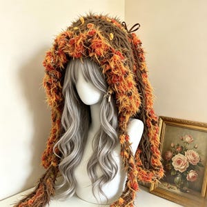 May include: A hooded accessory featuring a shaggy, textured brown yarn base with vibrant orange and yellow accents throughout. The hood has long side panels and a small brown ribbon bow at the top, displayed on a mannequin with long wavy silver hair.