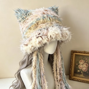 May include: A fuzzy, multi-colored bucket hat featuring cat-like ears and long, dangling braided tassels. The hat is crafted from textured, shaggy yarn in shades of cream, pale blue, and soft pink, accented with small, delicate ribbon bows.