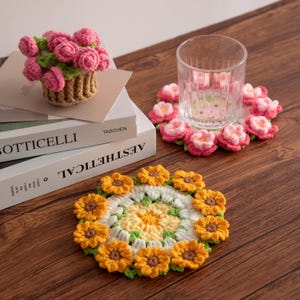 May include: Crocheted floral coasters and a small basket of pink roses. The coasters feature yellow sunflowers and pink flowers. A clear glass sits on a coaster with pink flowers. The items are on a wooden surface.