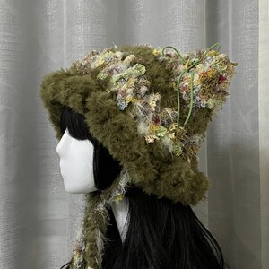 Puede incluir: Un sombrero verde oliva único con una apariencia texturizada y peluda. El sombrero presenta acentos florales coloridos y un lazo verde. El sombrero está diseñado con orejeras y una extensión tipo bufanda.